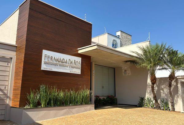 Fachada moderna da clínica odontológica Fernanda Da Ros, especializada em Odontologia Estética e Implantes, localizada em Leme-SP. A entrada conta com revestimento de madeira, placa de identificação, jardim com plantas ornamentais e palmeiras, além de um amplo espaço de estacionamento.
